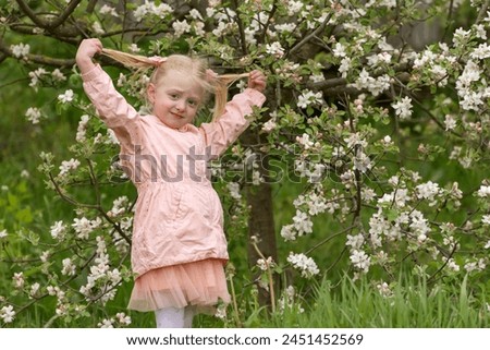 Cute blonde girl in pink jacket and skirt stands in blooming garden and holds herself by the tails. Spring garden.