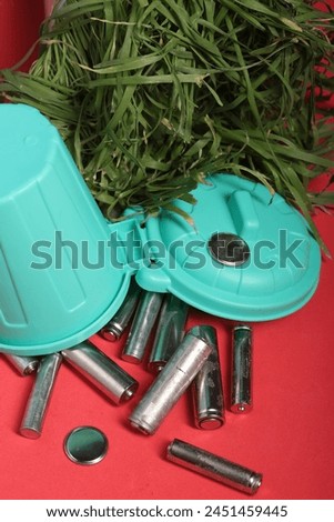 Used finger batteries in a miniature trash can. Fading greens. Pink background. 