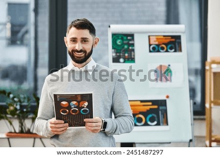 A man with beard smiling and holding charts while standing in office