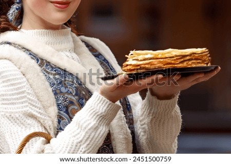 A young woman is dressed in national Russian dress. She is holding a plate of pancakes and a wicker basket in her hands. Greets guests for Maslenitsa