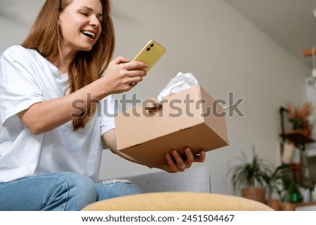 Joyful woman unpacking online purchase and taking picture of goods in cardboard. Online photo messaging, sharing picture in the internet. 