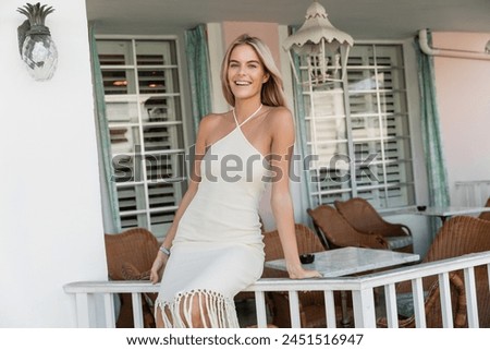 A young, beautiful blonde woman in a flowing white dress standing gracefully on a porch against a backdrop of the Miami skyline.