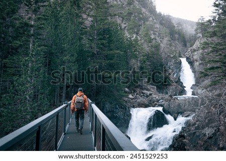 A man with an orange jacket and a backpack walks on an iron bridge. There is a waterfall near him. Concept of adventure and exploration. The man is hiking through nature.