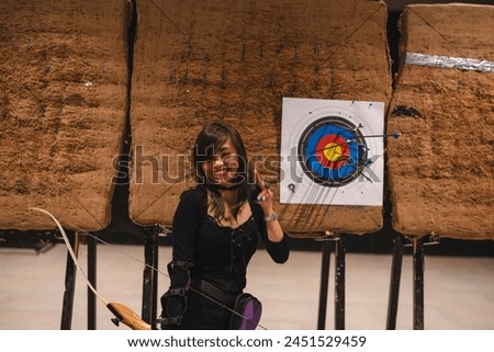 A cheerful young Asian female archer poses with her bow at an indoor archery range, dressed in casual attire and equipped with an arm guard, demonstrating her shooting results with evident pride. Royalty-Free Stock Photo #2451529459