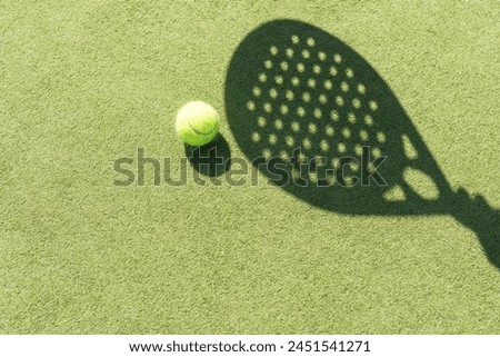 Shadow of a padel racket with a yellow ball on the green grass.