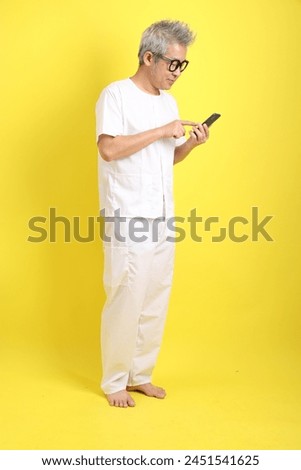 Chinese Vegetarian Festival. Senior man wearing white clothing with gesture of Holding mobile phone isolated on yellow background. Nine emperor god, J festival.