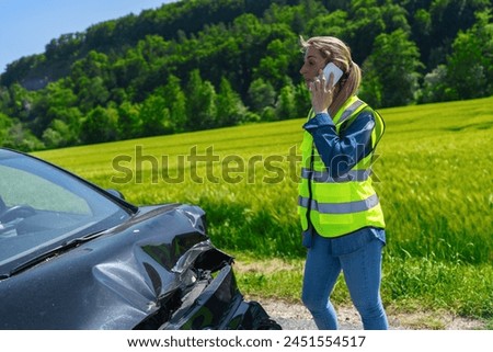 Woman in high visibility vest on phone with damaged car in foreground showing accident site on rural road Royalty-Free Stock Photo #2451554517