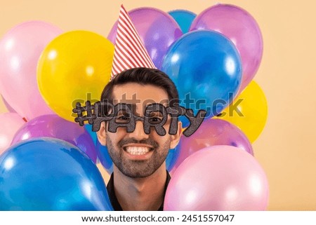 Close-up portrait of young man with party eye glasses and party hat looking at camera with smile amidst colourful balloons