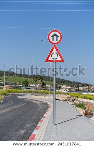 An urban area entrance sign coupled with a roundabout warning, under the clear spring sky of Israel.
