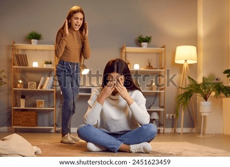 Angry child girl quarrels screams at mother who sits on floor, covers face in despair, thinks of disagreement, argument, misbehavior, scandal, hysterical tantrum caused by upbringing mistake failure Royalty-Free Stock Photo #2451623459