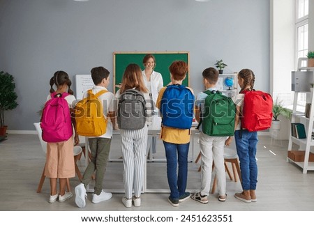 Small group of children in casual clothes gets acquainted with smiling teacher of individual classes. Teenagers with backpacks on their backs lined up in front of their teacher in the classroom. Royalty-Free Stock Photo #2451623551