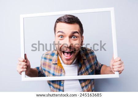 Crazy picture. Handsome young man in shirt holding picture frame in front of his face and smiling while standing against grey background 