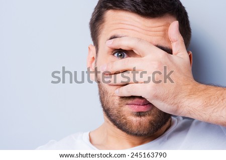 Shocked and terrified. Portrait of young man covering his face by hand and looking at camera while standing against grey background Royalty-Free Stock Photo #245163790