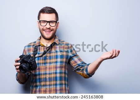 Come to me to get good photo. Portrait of confident young man in shirt holding camera and pointing away while standing against grey background