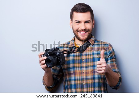 Good smile!. Portrait of confident young man in shirt holding camera and showing thumb up while standing against grey background