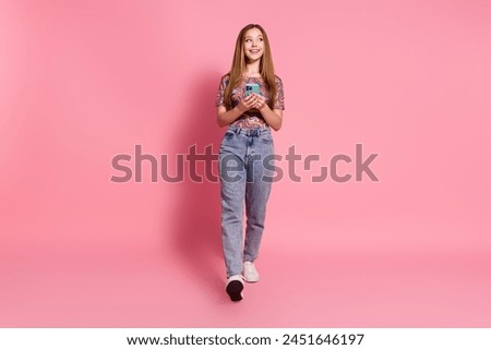 Full size photo of cheerful teen wear colorful t-shirt jeans hold smartphone look at offer empty space isolated on pink color background