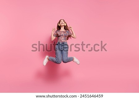Full length photo of lovely teen jump headphones point look up empty space dressed stylish print clothes isolated on pink color background