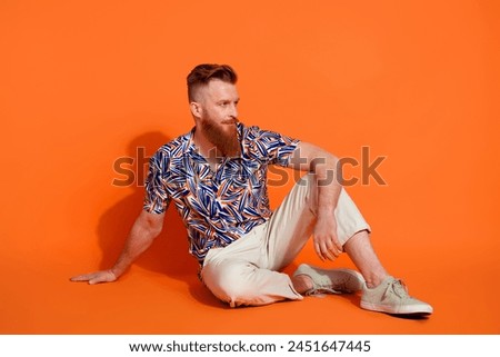 Full length photo of brutal guy wear print shirt trousers sit on floor look at offer empty space isolated on orange color background