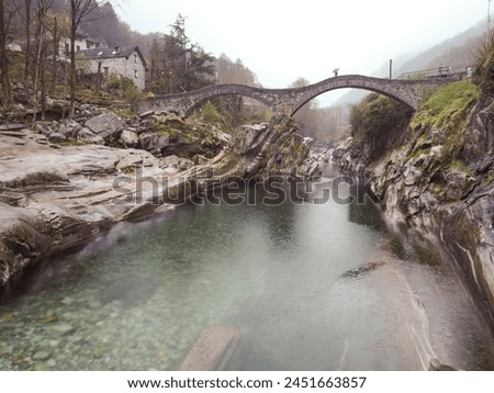A photo of a stone bridge over a river in a mountainous area. The bridge is covered in greenery and the river below is flowing through a rocky gorge. The background consists of a foggy mountain