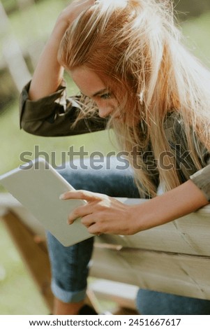 Young woman focused on digital tablet in hand, leaning on wooden bench outdoors
