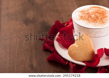 Lovely moment. Closeup image of heart shape cookie and cup of coffee placed with copy space on wooden table and decorated with rose petals
