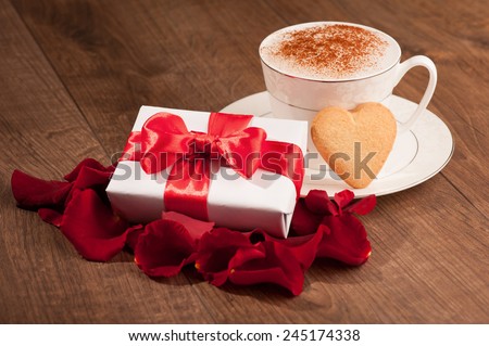 With love. Closeup image of cup of coffee with lovely heart shape cookie placed with a gift box on wooden table and decorated with rose petals
