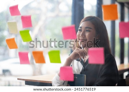 Smiling Asian businesswoman using colorful sticky notes for brainstorming on a glass wall in a modern office.

