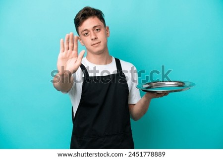 Young waitress with tray isolated on blue background making stop gesture