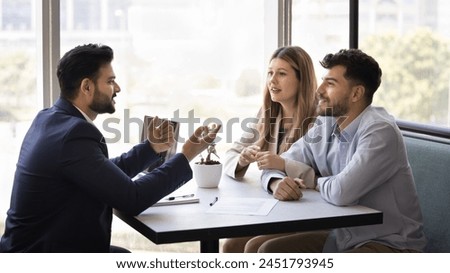 Positive married couple consulting financial expert, real estate agent at meeting, listening to young Indian business consultant in formal suit, sitting at table with paper agreement Royalty-Free Stock Photo #2451793945
