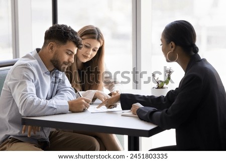 Confident young African realtor agent pointing at paper agreement for couple of customers, clients signing, giving consultation, legal help with mortgage, real estate property buying