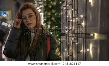 Beautiful young female model with glasses being outdoor in the street and standing next to the wall with hanging decorative lights.
