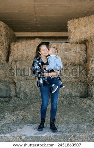Little girl sits in her mother arms and kisses her on the cheek among the bales of hay