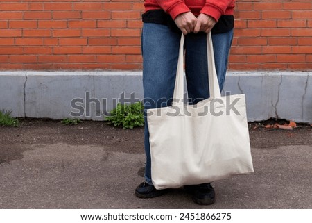 Woman in red hoodie holding tote canvas blank eco bag on street wall background. Female consumer hold white textile shopper. template or place for your design, logo, text