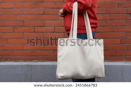 Woman in red hoodie holding tote canvas blank eco bag on street wall background. Female consumer hold white textile shopper. template or place for your design, logo, text