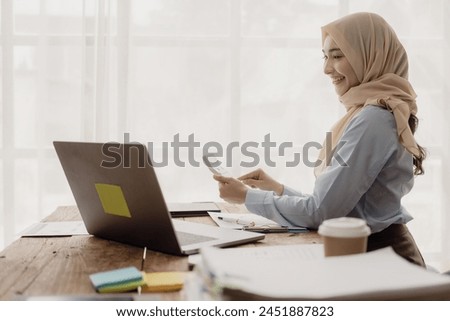 Asian Muslim businesswoman uses calculator and writes notes with mathematical financial calculations on table at office with laptop and financial documents, taxes, accounting, financial