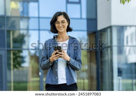 Confident young businesswoman with glasses using a smartphone outside a modern office building. This image captures a casual yet professional vibe.