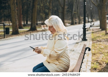 Elegant mature blonde woman sitting outdoors and holding smartphone gadget in hand for communicate on the city street. Urban, people concept. Spring sunny day