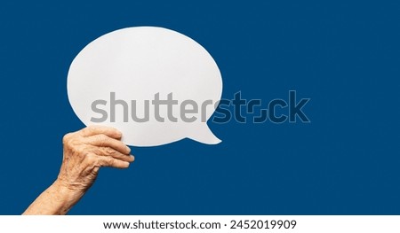 Close-up of a senior woman's hand holding a blank white speech bubble against a blue background. Space for text