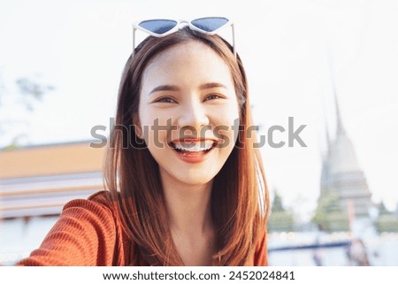 Happy young Asian woman traveler wearing orange shirt with taking selfie photo on camera at temple or palace wall. summer tourism concept.