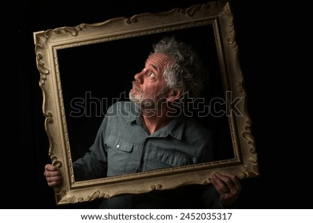 Black and white portrait of an attractive older Caucasian man with grey hair and beard. Turned to the upper right side, looking through a picture frame with a quizzical expression. 
