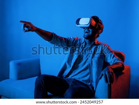 Man watching at funny movie by using virtual reality glass. Relaxed teenager enjoy watching movie and playing game while sitting at sofa with colorful neon light. Technology innovation. Deviation.