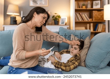 Young woman checking temperature with hand of her ill son. Mother checking temperature of her sick boy. Sick child lying on bed under blanket with woman checking fever on forehead by hand.