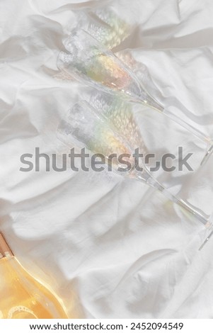 Rainbow color shining champagne glasses and white sparkling wine bottle on bed, on white blanket background. Lifestyle aesthetic photo, star filter effect. Romance meeting, romantic holiday concept. 