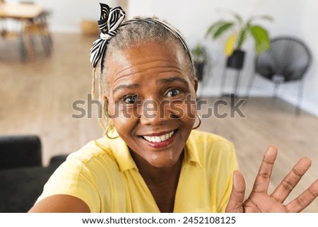 African American senior woman showing peace sign at home during a video call, wearing a yellow shirt. She has short gray hair, light wrinkles, and a bright smile, unaltered.