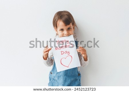 little cute white brunette girl 1.5 years old holds in her hands and teeth a card with the inscription text happy mothers day and a red heart on a white background