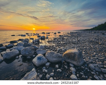 Sunset over serene lakeshore, with calm water reflecting the blue sky. Moss-covered rocks of varying sizes and shapes add texture, while the warm glow of the setting sun creates a tranquil atmosphere. Royalty-Free Stock Photo #2452161187