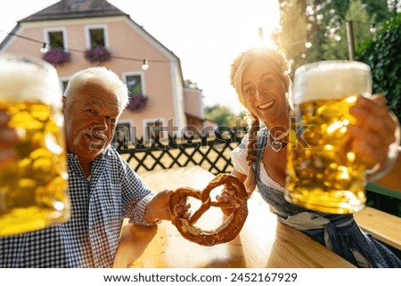 Senior man and young woman in Bavarian tracht holding beer mugs and pretzel, smiling at camera in a beer garden or Oktoberfest, Munich, Germany Royalty-Free Stock Photo #2452167929