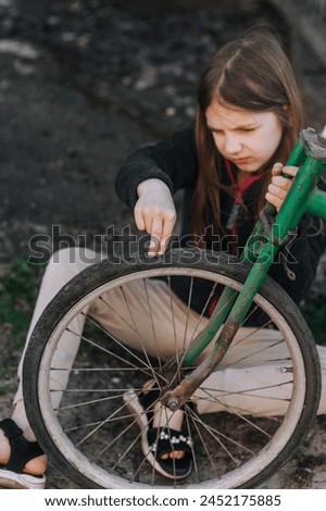 Little sad teenage girl, dissatisfied distressed child sitting near an old bicycle with a broken, punctured wheel tire outdoors. Photography, portrait. Royalty-Free Stock Photo #2452175885