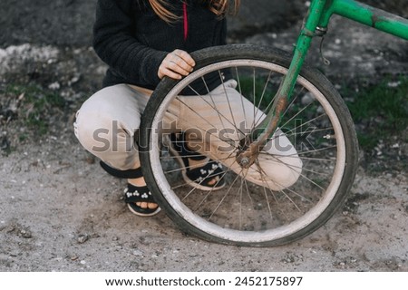 Little teenage girl, dissatisfied, distressed child sits near an old bicycle with a broken, punctured wheel tire outdoors. Photography, portrait. Royalty-Free Stock Photo #2452175897