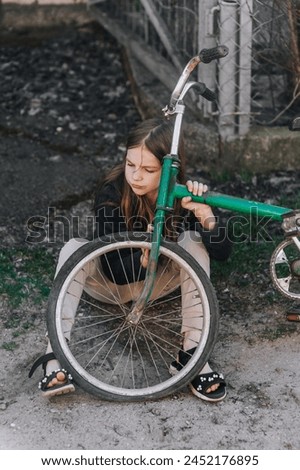 Little teenage girl, dissatisfied, distressed child sits near an old bicycle with a broken, punctured wheel tire outdoors. Royalty-Free Stock Photo #2452176895